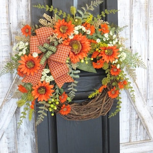 May include: A decorative wreath featuring bright orange sunflowers, small white flowers, and green fern foliage arranged on a natural brown twig base. An orange and white checkered ribbon bow with a green accent is tied to the side of the wreath.