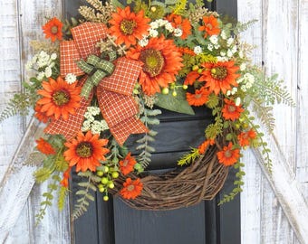 May include: A decorative wreath featuring bright orange sunflowers, small white flowers, and green fern foliage arranged on a natural brown twig base. An orange and white checkered ribbon bow with a green accent is tied to the side of the wreath.