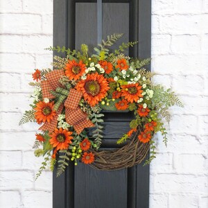 May include: A decorative wreath featuring vibrant orange sunflowers, small white filler flowers, and green fern foliage. A plaid ribbon in shades of orange and green is tied to the side. The base is constructed from natural brown woven twigs.