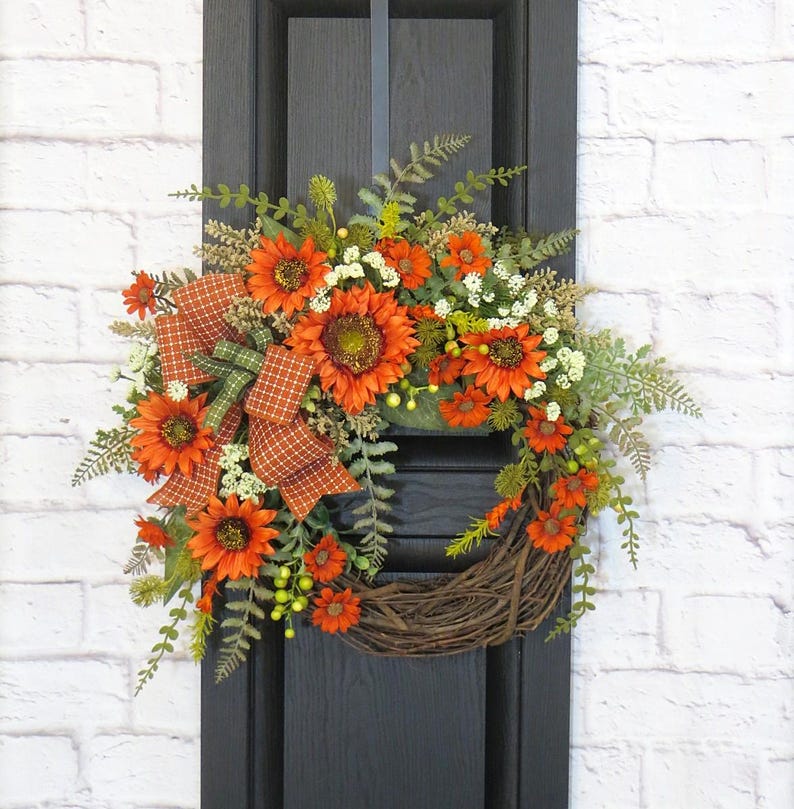 May include: A decorative wreath featuring vibrant orange sunflowers, small white filler flowers, and green fern foliage. A plaid ribbon in shades of orange and green is tied to the side. The base is constructed from natural brown woven twigs.