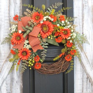 May include: A decorative wreath featuring vibrant orange sunflowers, small white filler flowers, and green fern foliage arranged on a circular brown twig base. An orange and white checkered ribbon with a green accent bow is tied to the side.