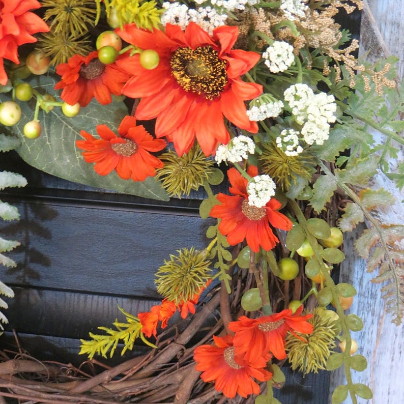 May include: A decorative wreath featuring vibrant orange sunflowers and smaller orange daisies, accented with clusters of small white flowers, green berries, and textured green foliage, all arranged on a base of intertwined brown natural twigs.