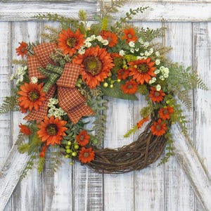 May include: A decorative wreath featuring orange sunflowers, small white flowers, and green foliage arranged on a circular brown twig base. A plaid ribbon in shades of orange, brown, and green is tied to the left side of the floral arrangement.