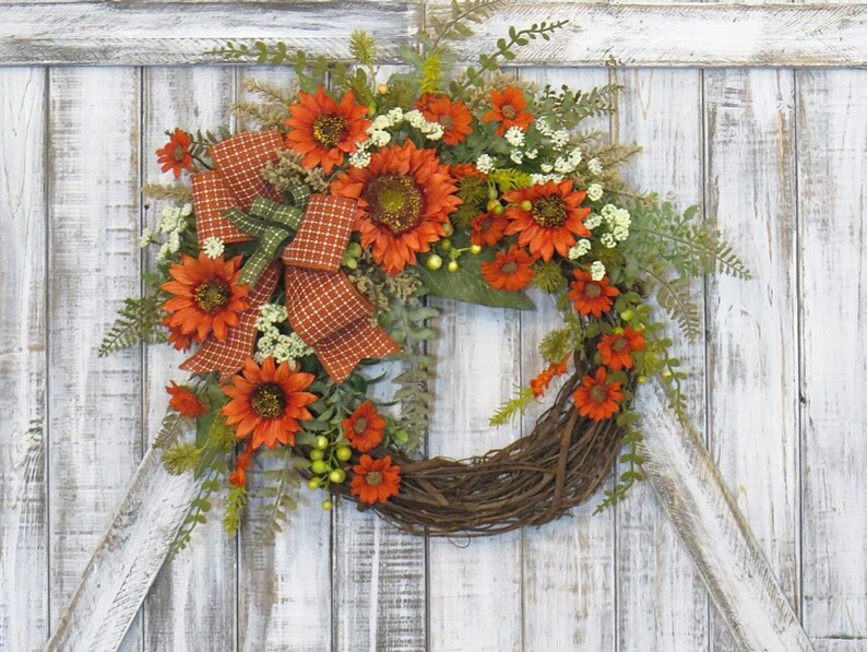 May include: A decorative wreath featuring orange sunflowers, small white flowers, and green foliage arranged on a circular brown twig base. A plaid ribbon in shades of orange, brown, and green is tied to the left side of the floral arrangement.