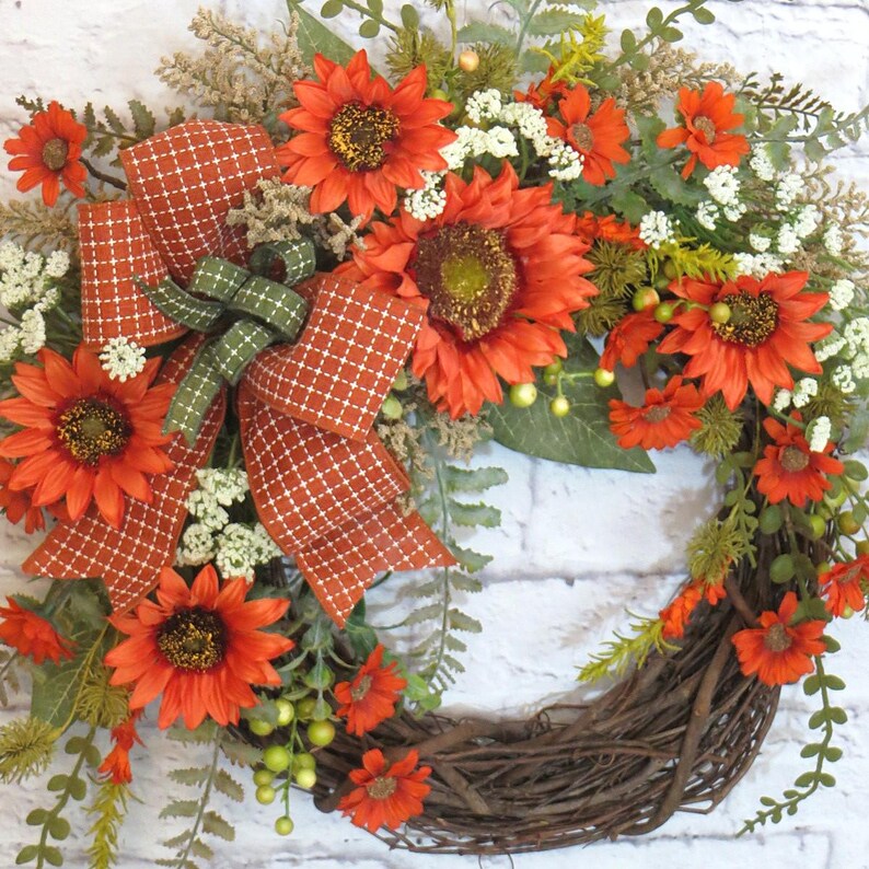 May include: A decorative wreath featuring vibrant orange sunflowers, small white filler flowers, and green foliage arranged on a natural twig base. A large, burnt orange bow with a white grid pattern is centered among the floral elements.