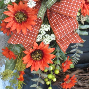May include: A decorative wreath featuring vibrant orange sunflowers, small white filler flowers, and green foliage. A large, textured orange ribbon with a white cross-stitch pattern is tied at the top, accented by a smaller green ribbon, all set on a twig base.
