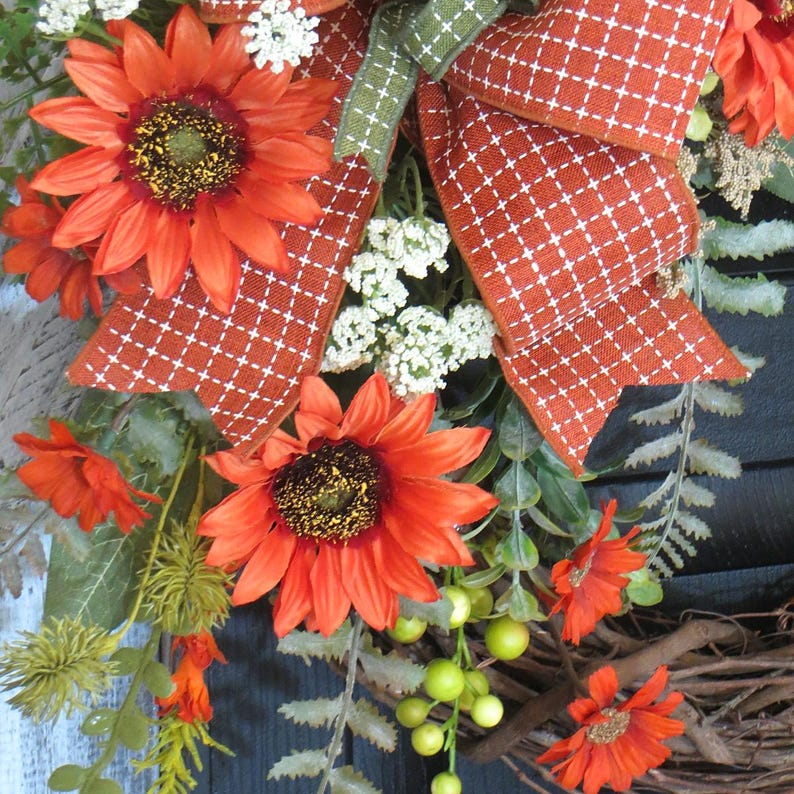 May include: A decorative wreath featuring vibrant orange sunflowers, small white filler flowers, and green foliage. A large, textured orange ribbon with a white cross-stitch pattern is tied at the top, accented by a smaller green ribbon, all set on a twig base.