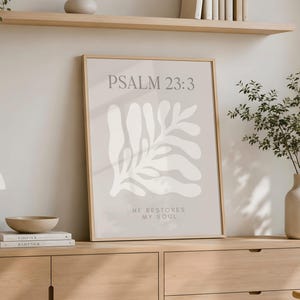 May include: A framed wall art print featuring the text PSALM 23:3 and HE RESTORES MY SOUL in dark gray lettering, centered above and below a large, abstract, off-white organic leaf shape on a light beige background within a light wood frame.