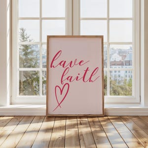 Könnte beinhalten: Ein gerahmter Druck mit den Worten "have faith" in einer Schreibschrift und einem Herzsymbol, alles in leuchtendem Pink. Das Kunstwerk ist vor einem zartrosa Hintergrund platziert und in hellem Holz gerahmt. Der Druck lehnt an einem Fenster.