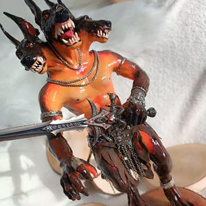 Könnte beinhalten: Detaillierte Figur einer dreiköpfigen hundeartigen Kreatur mit muskulösem Oberkörper, die ein Schwert hält. Die Figur hat einen rot-orangen Hautton mit schwarzen und roten Akzenten. Sie ist mit Silberschmuck und Ketten verziert und hat scharfe Zähne.