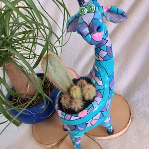 Könnte beinhalten: Ein handbemalter Giraffen-Blumentopf in Blau-, Lila- und Rosatönen, der Kakteen hält. Die Giraffe ist mit mosaikartigen Mustern verziert und steht auf Holzscheiben. Im Hintergrund befindet sich ein blauer Topf mit einer Pflanze.