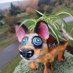 Könnte beinhalten: Handbemalter Hund-Blumentopf in Orange, Schwarz und Weiß. Der Hund hat große blaue Augen und rosa Ohren. Der Topf enthält eine grüne Pflanze mit langen, schmalen Blättern. Der Pflanztopf steht auf einer strukturierten, grünen Oberfläche.