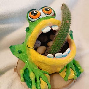 Könnte beinhalten: Ein skurriler, handbemalter Keramik-Frosch-Pflanztopf mit weit geöffnetem Maul, das einen kleinen Kaktus enthält. Der Frosch ist überwiegend grün und gelb, mit orangefarbenen Augen und einem Holzsockel. Der Kaktus ist grün und stachelig.