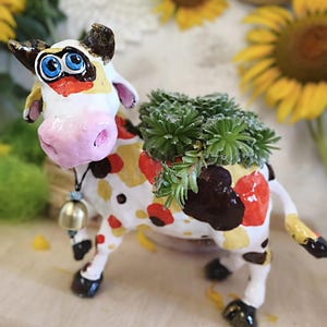 Könnte beinhalten: Ein kuhförmiger Pflanztopf aus Keramik mit weißem Körper und braunen, gelben sowie roten Flecken. Die Kuh hat große blaue Augen, eine rosa Schnauze und eine kleine Metallglocke um den Hals. Eine grüne Sukkulente wächst auf dem Rücken der Kuh.