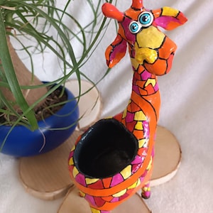 Könnte beinhalten: Ein farbenfroher, giraffe-förmiger Blumentopf mit orangefarbenem Körper und Mosaikmustern in Gelb, Pink und Rot. Die Giraffe hat blaue Augen und einen schwarzen Innenraum. Sie steht auf Holzscheiben.