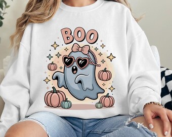 Könnte beinhalten: Ein weißes Langarm-Sweatshirt mit einer Grafik eines hellblauen Geistes, der eine herzförmige Sonnenbrille und eine Schleife trägt. Der Geist ist von kleinen Kürbissen und Sternen umgeben, darüber steht der Text BOO in großen Buchstaben gedruckt.
