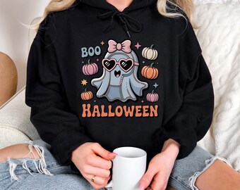 Könnte beinhalten: Ein schwarzer Kapuzenpullover mit einer Grafik eines hellblauen Geistes, der eine herzförmige Sonnenbrille und eine rosa Schleife trägt. Das Design enthält den Text BOO und HALLOWEEN, umgeben von kleinen orangefarbenen, rosa, weißen Kürbissen und Sternen.