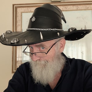 May include: A black leather witch hat featuring a pointed crown, a wide brim, and a silver-toned chain band. The hat is decorated with a small circular pentagram charm on the front, along with several silver-colored metal spikes and skull accents.