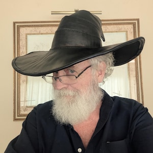 May include: A black leather hat with a wide, curved brim and a tall, slightly pointed crown. The hat features visible stitched seams along the crown and the edge of the brim, creating a structured appearance with a smooth, dark finish.