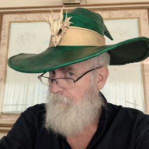 May include: A wide-brimmed green hat featuring a tan leather band around the crown and a cream-colored dragon skull ornament attached to the front. The hat has a pointed top and a structured brim with visible stitching along the edges.