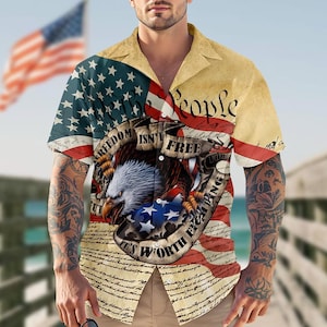 Könnte beinhalten: Kurzärmeliges Button-Down-Hemd mit einem Design der amerikanischen Flagge, einem Adler, Sternen und Streifen. Das Hemd enthält den Text "Freedom Isn't Free, It's Worth Fighting For". Das Hemd hat einen beigefarbenen Kragen und ein patriotisches Thema.