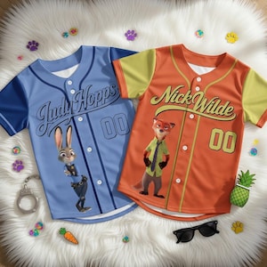 Könnte beinhalten: Zwei Baseball-Trikots, eines hellblau und eines orange, mit Charakteren aus Zootopia. Das hellblaue Trikot trägt die Aufschrift "Judy Hopps" und das orangefarbene Trikot "Nick Wilde". Beide Trikots haben die Nummer "00".