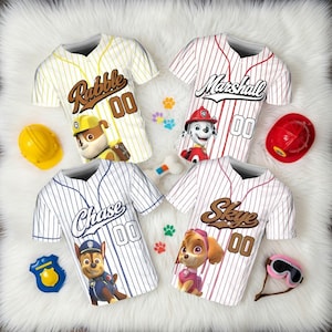 Könnte beinhalten: Vier Baseball-Trikots mit Paw Patrol-Charakteren: Rubble, Marshall, Chase und Skye. Jedes Trikot hat ein Charakterbild, Namen und die Nummer 00. Zubehör umfasst einen gelben Helm, einen roten Helm, eine Plakette, einen Knochen und eine rosa Brille.
