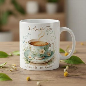 Könnte beinhalten: Weißer Keramikbecher mit Henkel, mit einer Aquarellillustration einer Teetasse und Untertasse. Die Teetasse ist mit Tee gefüllt und hat Dampf. Der Becher hat den Text "I Am the Tea" und "I am the are having".