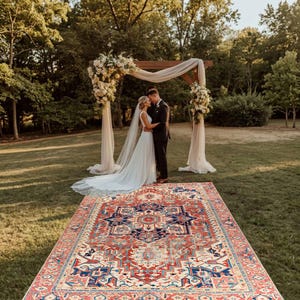 Puede incluir: Una ceremonia de boda al aire libre con una pareja abrazándose bajo un arco de madera decorado con flores y tela drapeada. Una alfombra estampada conduce al arco. La novia lleva un vestido blanco y velo, y el novio un traje oscuro.