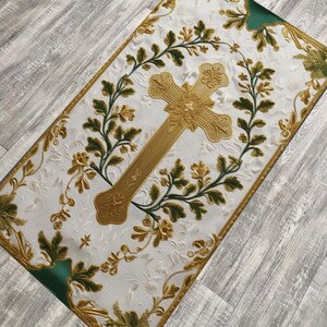 May include: A rectangular white textile with a gold cross at the center, surrounded by a wreath of green and gold floral designs. The edges are trimmed with gold and green accents, creating a decorative religious item.