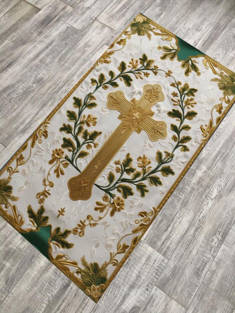 May include: A rectangular white textile with a gold cross at the center, surrounded by a wreath of green and gold floral designs. The edges are trimmed with gold and green accents, creating a decorative religious item.