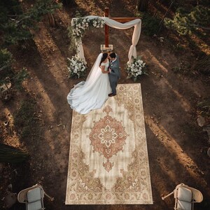 May include: An aerial view of a wedding ceremony in a woodland setting. A couple stands on a large, rectangular rug with an ornate, floral design in beige, red, and green. A wooden cross and floral archway are behind them.