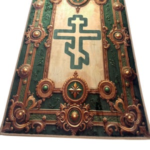 May include: A rectangular green and gold decorative rug with an ornate design. The rug features a large green cross in the center, surrounded by gold embellishments and a green border. The rug has a beige background.