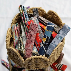 May include: A collection of fabric wristlet keychains in a woven straw basket. The wristlets feature various colourful patterns including floral, geometric, and architectural prints. Each strap is stitched with a small fabric label attached.