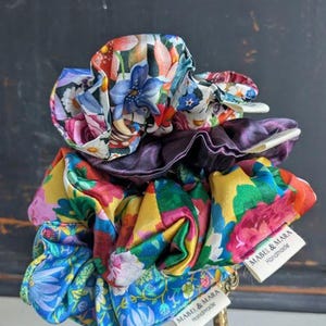 Op de afbeelding: Een stapel kleurrijke bloemen scrunchies. De scrunchies hebben verschillende patronen met bloemen in blauw, rood, geel en paars. Elke scrunchie is gemaakt van een zijdeachtige stof. Het label luidt "MABEL & MARA Handmade".