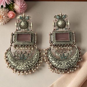 Può includere: Un paio di orecchini color argento con un centro rettangolare e una parte inferiore a forma di mezzaluna. Gli orecchini presentano dettagli intricati, tra cui piccole perle ed elementi decorativi. Gli orecchini sono progettati per orecchie forate.