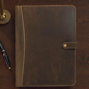 Puede incluir: Un portafolio de cuero marrón con un panel vertical contrastante en el lado izquierdo. El portafolio tiene un cierre de botón y está sobre una superficie de madera oscura. Un bolígrafo es visible a la izquierda y otro en la parte inferior.