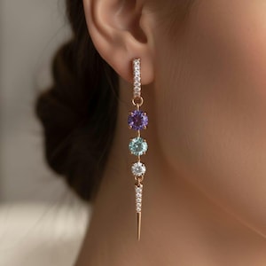 Könnte beinhalten: Ohrringe aus Roségold mit einem hängenden Design. Die Ohrringe zeigen eine Reihe farbiger Edelsteine: lila, hellblau und weiß. Der obere Teil des Ohrrings ist mit kleinen, klaren Steinen besetzt, und der untere Teil hat eine spitze Spitze.