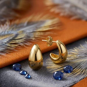 Könnte beinhalten: Ein Paar polierte Gold-Creolen mit glattem, tropfenförmigem Design. Die Ohrringe verfügen über einen Steckerverschluss und werden von fünf kleinen, facettierten, tiefblauen ovalen Edelsteinen begleitet, die auf der Oberfläche liegen.