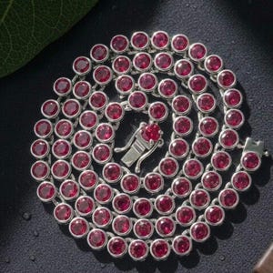 May include: A silver-toned chain necklace featuring a continuous series of round, faceted, deep red stones set in individual metal bezels. The necklace is coiled in a circular pattern and includes a secure metal box clasp closure at the center.