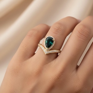 Könnte beinhalten: Ein goldener Ring mit einem tropfenförmigen, dunkelgrünen Edelstein, umgeben von kleinen, klaren Steinen. Das V-förmige Band ist ebenfalls mit kleinen, klaren Steinen besetzt. Der Ring befindet sich an einem Finger.