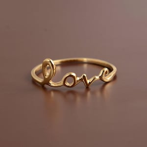 Puede incluir: Un anillo dorado que deletrea la palabra "love" en una fuente cursiva. El anillo es delgado y delicado, con las letras conectadas. El anillo probablemente está hecho de oro o un metal dorado.