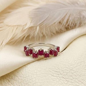 Könnte beinhalten: Ein silberner Ring mit einer Reihe quadratisch geschliffener, tiefroter Edelsteine. Der Ring ist vor einem weichen, weißen Hintergrund mit einer Feder in der oberen linken Ecke platziert. Die Edelsteine sind gleichmäßig angeordnet.