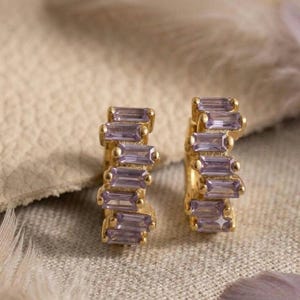 May include: A pair of gold hoop earrings featuring a row of rectangular, baguette-cut lavender stones set along the front. The earrings have a polished gold finish and a curved design that showcases the light purple gemstones in a vertical arrangement.
