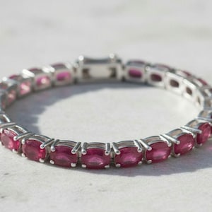 Op de afbeelding: Een zilverkleurige armband met een doorlopende rij ovaal geslepen, diep roze-rode edelstenen. Elke steen is individueel gezet in een metalen zetting met vier pootjes, verbonden door kleine schakels en voorzien van een rechthoekige sluiting.