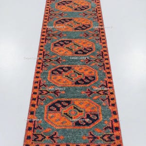 May include: A rectangular runner rug with a teal background and an intricate pattern in orange, black, and red. The design features repeating geometric shapes and borders. The rug is likely made of woven material.