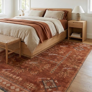 May include: A bedroom scene featuring a bed with beige bedding and a rust-colored throw blanket. A patterned rust-colored rug covers the floor. A wooden nightstand with a lamp and a woven bench are also visible.