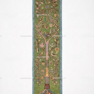 Könnte beinhalten: Ein rechteckiger grüner Teppich mit einem Baumdesign. Der Baum zeigt Vögel, Früchte und einen karierten Stamm. Der Rand ist blau und der Teppich hat einen Fransenrand. Das Design ist detailliert und farbenfroh.
