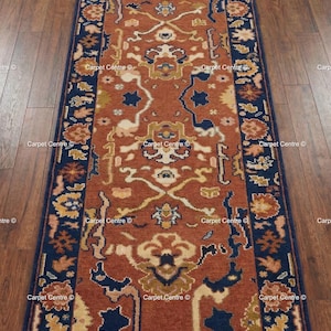 May include: A rectangular, ornate rug with a central rust-colored field and a navy blue border. The rug features intricate floral and geometric patterns in shades of beige, gold, and navy. The rug is placed on a dark wood floor.