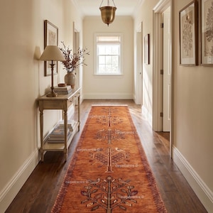 May include: An orange runner rug with a tree of life design, placed in a hallway with dark wood flooring. The rug features a repeating pattern of trees and birds, with a fringed edge. The walls are a light beige color.